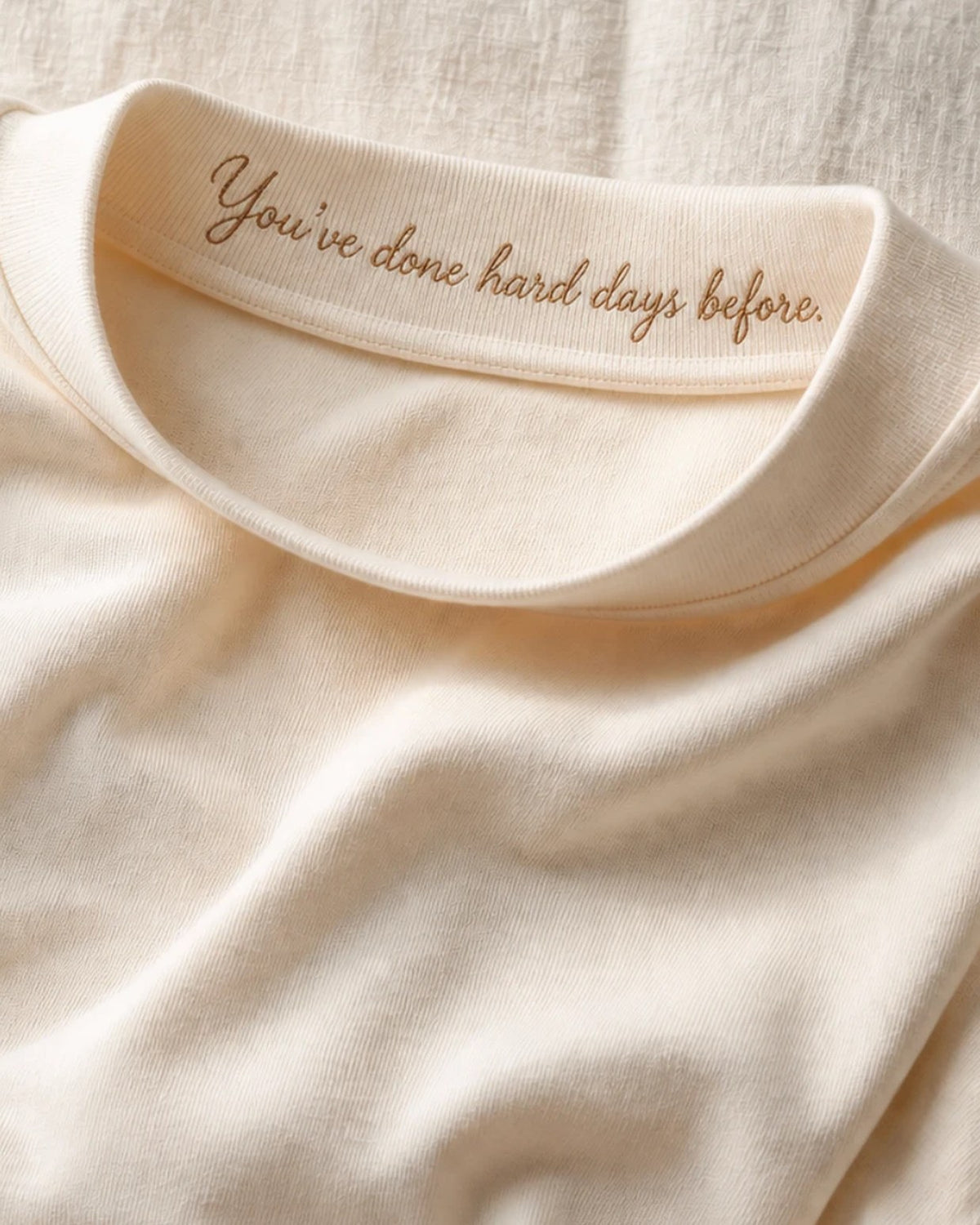 Close on the Cream collar band, tipped forward: 'You've done hard days before.' embroidered in cursive on the inner face, the thread raised above the cloth.