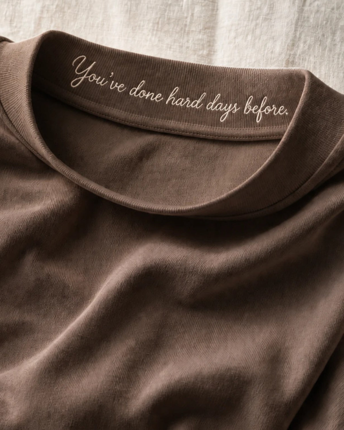 Close on the Mocha collar band, tipped forward: 'You've done hard days before.' embroidered in cream thread on the inner face, raised above the cloth.