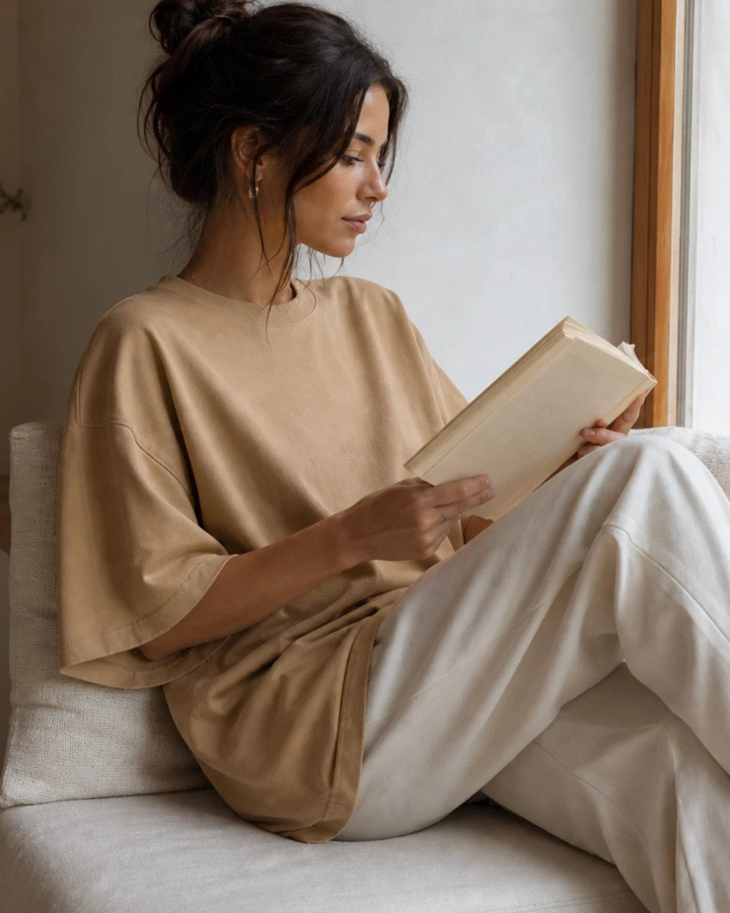 The Sand tee worn reading in a window seat, the oversized sleeve pushed back off the forearm.
