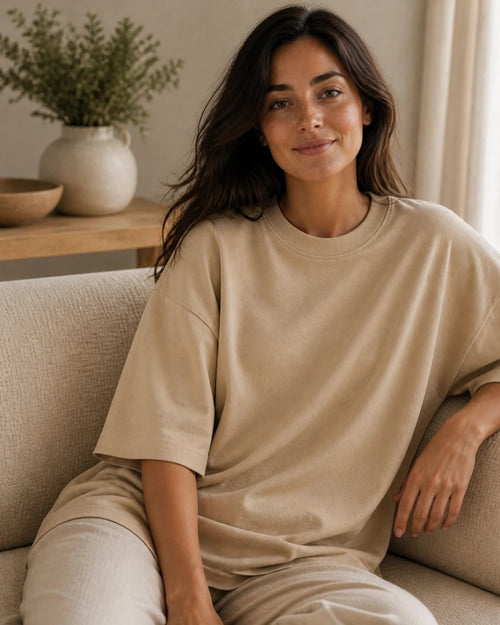 The oversized tee in Sand, worn loose on a sofa. Nothing is printed on the outside.